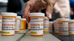 A round orange bottle of the drug oxycontin with the label carrying the expiration date and the website of the pharmacy. A hand seems to be picking up the bottle in the main frame, with several other bottles lined neatly behind them.